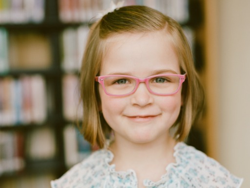Looking smart a young blue eyed girl with pink glasses smiles. The books in the background slightly blurred blend the ambiance for happiness that comes from learning.